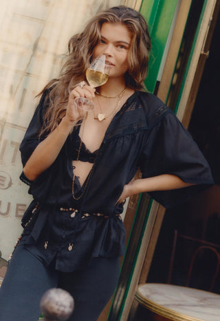 A woman with long wavy brown hair wearing a black bohemian-style sheer blouse and matching pants, holding a glass of white wine in front of her face. She is posed in front of a rustic doorway.