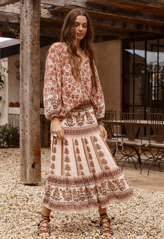 A woman wearing a bohemian-style two-piece dress set with a rust brown and cream floral print, standing on a gravel patio in front of a rustic wooden structure.